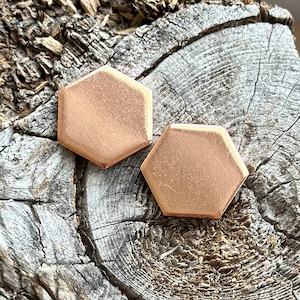 May include: Two hexagonal shaped earrings made of rose gold metal. The earrings are sitting on a piece of weathered wood.