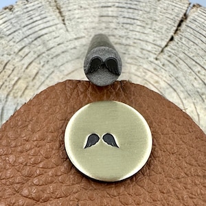 Op de afbeelding: Een metalen stempel met een vleugelontwerp is geplaatst boven een kleine, ronde, goudkleurige metalen schijf met hetzelfde vleugelontwerp. De schijf ligt op een bruin leren oppervlak, tegen een houten achtergrond met textuur.