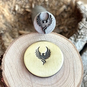 Könnte beinhalten: Nahaufnahme eines Stempels aus Metall mit einem Phönix-Motiv. Der Stempel liegt auf einer Holzoberfläche. Der Phönix ist ein mythischer Vogel, der Wiedergeburt und Erneuerung symbolisiert.