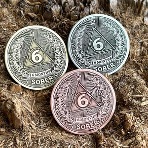 Peut inclure: Trois pièces de monnaie en métal ton argent, or et cuivre avec un motif triangulaire et le chiffre 6 au centre. Chaque pièce porte l'inscription "6 Months Sober" sur le bord.