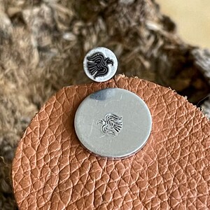 May include: Two silver metal buttons with a black and white bird design. One button is small and round, the other is larger and circular. They are placed on a brown leather surface.