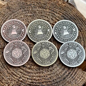 Op de afbeelding: Zes metalen tokens met verschillende ontwerpen. Drie tokens hebben een bed met een wassende maan en sterren, met de tekst "Go to Sleep" rond de rand. Drie tokens hebben een 20-zijdige dobbelsteen met de tekst "One More Game" rond de rand.