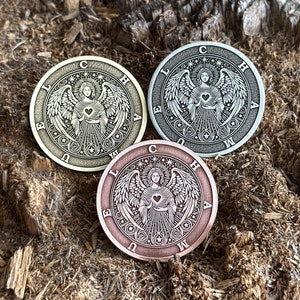 Puede incluir: Tres fichas metálicas redondas con un diseño de ángel. Cada ficha tiene un color diferente: oro, plata y cobre. El ángel sostiene un corazón en sus manos. Las fichas tienen las letras "E U L C H A M" alrededor del borde.