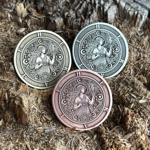 May include: Three circular metal coins featuring an engraved design of a woman holding a torch, encircled by text and stars. The coins are in bronze, silver, and gold tones, set on a textured wooden surface.