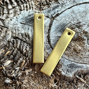 Puede incluir: Dos etiquetas rectangulares de metal de color dorado con un agujero en la parte superior. Las etiquetas están sobre una superficie de madera marrón rugosa.