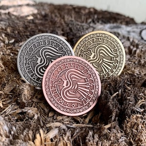May include: Three round metal coins with a stylized raven design and runes around the edge. The coins are in silver, gold, and copper.