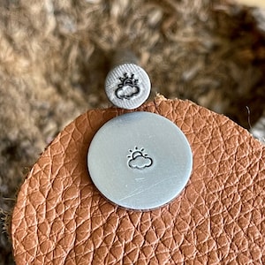 May include: A close-up of a silver metal stamp with a design of a sun and a cloud. The stamp is on a brown leather surface.