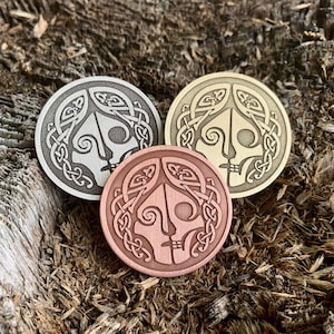 May include: Three circular metal pins with a Celtic knot design and a stylized face in the center. The pins are in silver, gold, and copper.