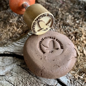 May include: A close-up of a brown clay disc with a leaf design stamped into it. A brass stamp with a bird design is visible in the background.