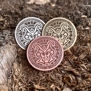 May include: Three round metal coins with a raised design of a jester's face with horns and runes around the edge. The coins are in silver, gold, and copper.
