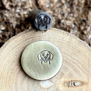 Könnte beinhalten: Ein Metallstempel mit einem Hundegesicht-Design. Der Stempel besteht aus Metall und hat einen Griff. Das gestempelte Bild ist eine detaillierte Umrisszeichnung eines Hundegesichts, mit herausgestreckter Zunge. Der Stempel wird auf einer Holzoberfläche gezeigt.