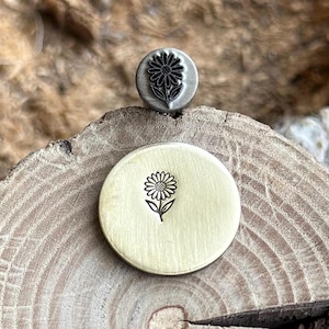 May include: A metal stamp and its impression. The stamp is round with a daisy design. The impression is on a round, gold-colored metal disc. The daisy design is black. The stamp is silver-colored.