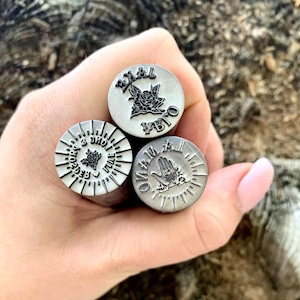 Może przedstawiać: Trzy metalowe stemple z grawerowanymi wzorami. Jeden stempel przedstawia różę i tekst, drugi ma wzór promieni słonecznych z tekstem i kwiatem, a trzeci przedstawia dłoń z okiem i tekstem. Stemple są trzymane w dłoni.