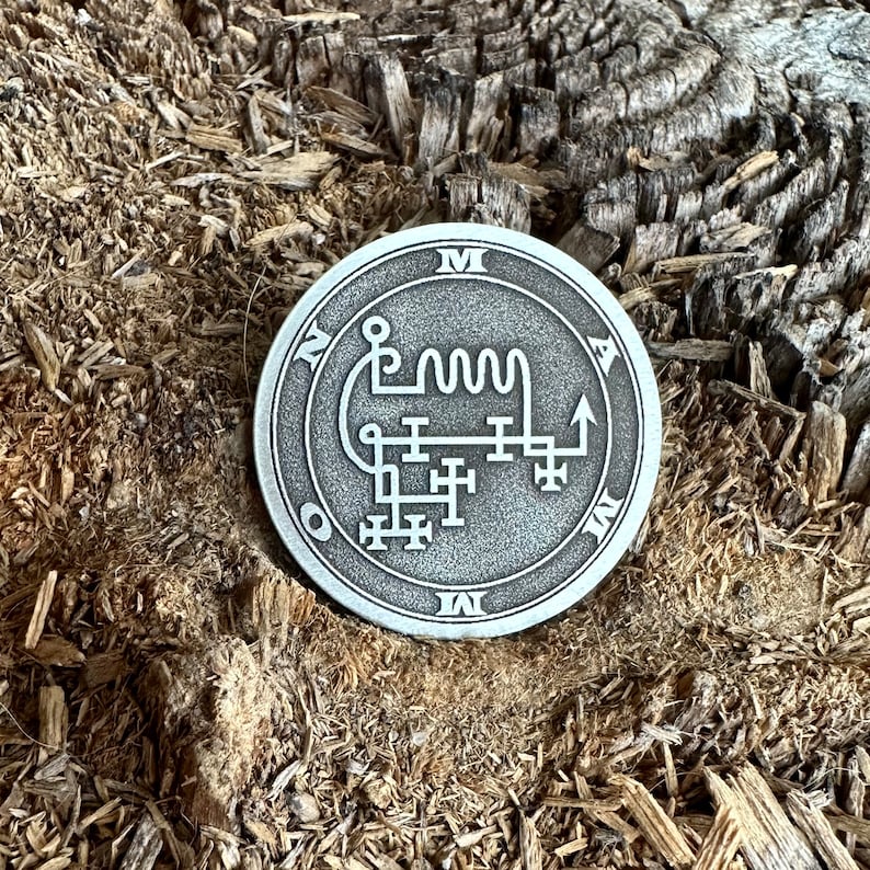 May include: A silver-toned metal pendant with a sigil inscribed on it. The sigil is a complex design of lines and symbols, including a wavy line and an arrow pointing upwards. The pendant is surrounded by a circle with the letters "M", "A", "O", and "I" inscribed around it.