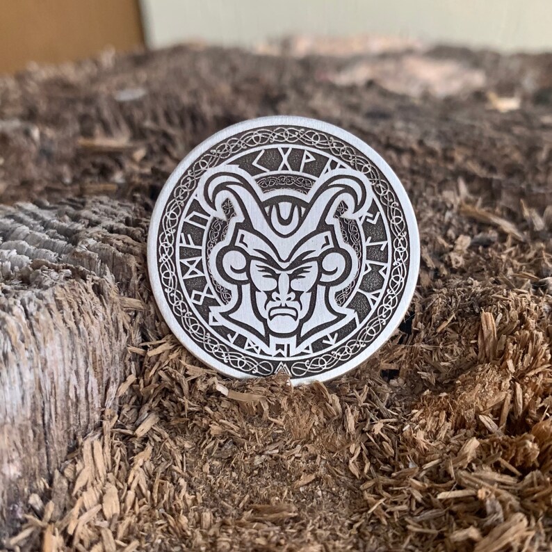 May include: A silver-toned metal coin with an intricate design featuring a stylized face with horns and a crown. The coin has a circular border with the text "XP" and other symbols.