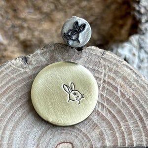 以下が含まれることがあります： ウサギのデザインの金属スタンプが2つ。1つは、ウサギの画像が浮き彫りになった、小さくて丸い灰色のスタンプです。もう1つは、ウサギの画像が彫り込まれた、大きくて丸い金色のスタンプです。スタンプは木製の表面にあります。