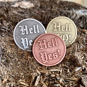 May include: Three round metal coins with the phrase "Hell Yes!" in a gothic font. The coins are in silver, gold, and copper.