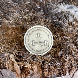 May include: A circular, metallic coin with an engraved design. The coin features a central symbol with three crosses and a curved line, surrounded by a circular border with the letters "H" and "L". The coin is set against a textured, wooden background.