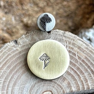 Może przedstawiać: Dwa metalowe stemple, jeden okrągły i jeden okrągły, każdy ze szczegółowym wzorem kwiatowym. Górny stempel jest szary, a dolny złoty. Wzór kwiatowy jest wytrawiony na czarno, ukazując szczegóły płatków i łodygi.