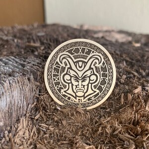 May include: A circular wooden token with a detailed engraving of a face with horns and a crown. The face has a serious expression and is surrounded by a circular pattern of runes. The text "XP" is visible on the top of the token.