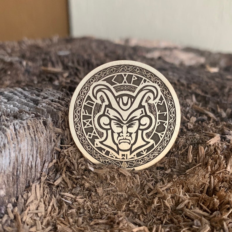 May include: A circular wooden token with a detailed engraving of a face with horns and a crown. The face has a serious expression and is surrounded by a circular pattern of runes. The text "XP" is visible on the top of the token.