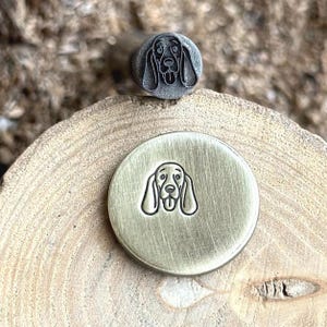 May include: Two metal stamps with a dog face design. One stamp is a small, round stamp with a detailed dog face. The other stamp is a larger, round stamp with a simplified dog face design. The stamps are on a wooden surface.