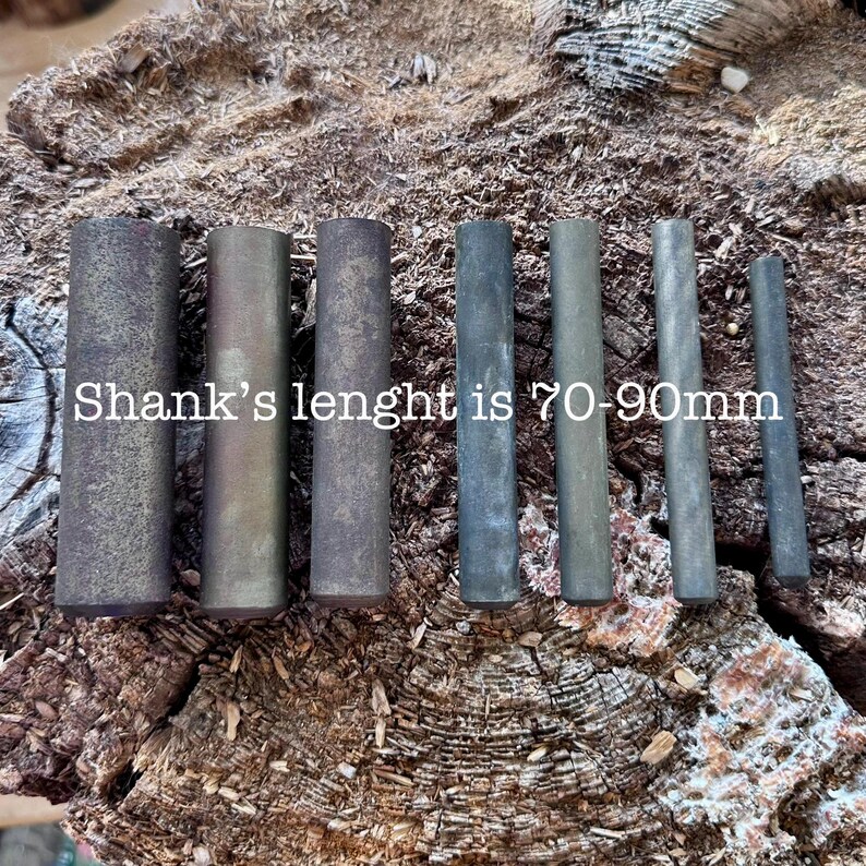 May include: A collection of cylindrical metal rods in varying shades of gray and brown, arranged on a textured wooden surface. The text "Shank's lenght is 70-90mm" is visible. The rods appear to be of different lengths and diameters.