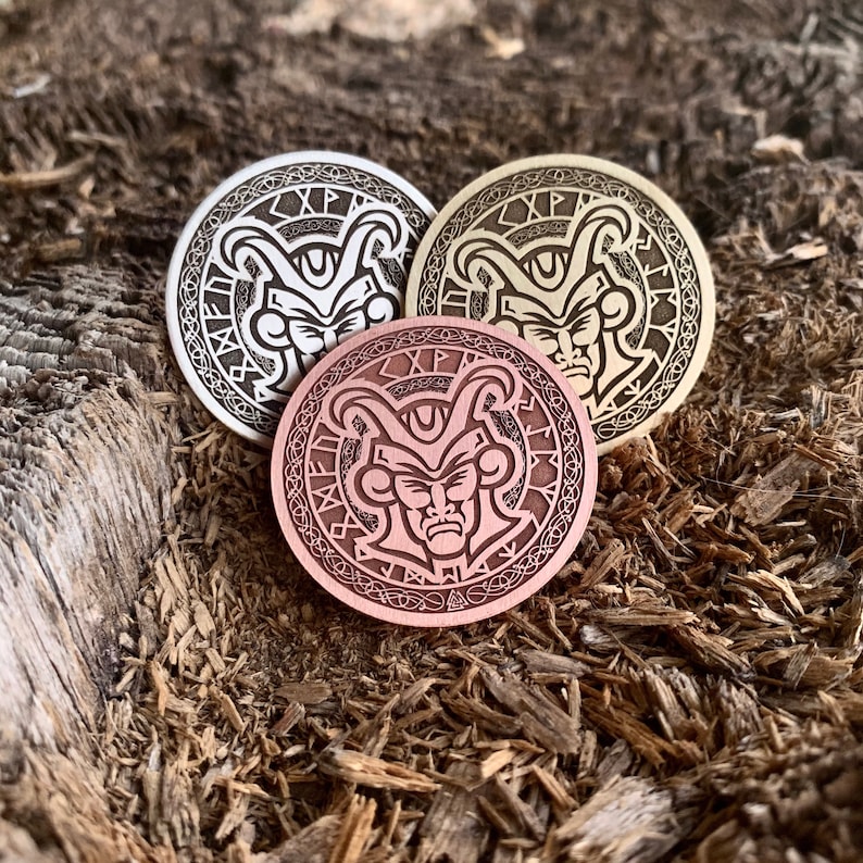 May include: Three round metal coins with a raised design of a jester's face with horns and runes around the edge. The coins are in silver, gold, and copper.