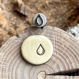 Könnte beinhalten: Zwei Metallstempel mit einem Tropfendesign. Ein Stempel ist silberfarben mit schwarzer Tropfenkontur, der andere goldfarben mit Tropfenkontur. Die Stempel sind kreisförmig und liegen auf einer Holzoberfläche.