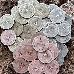 Può includere: Una collezione di gettoni di sobrietà rotondi in metallo di vari colori, tra cui oro, argento e rame. Ogni gettone presenta un design triangolare con un numero che rappresenta la durata della sobrietà, come 1 mese, 3 mesi, 6 mesi, 11 mesi e 365 giorni. I gettoni sono disposti in una pila su una superficie marrone testurizzata.
