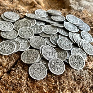 以下が含まれることがあります： 複雑なデザインが施された銀色の金属製トークンのコレクション。各トークンには、ユニークなシンボルまたは紋章が描かれています。