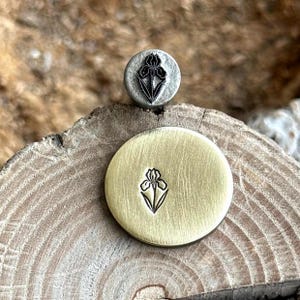 Op de afbeelding: Twee metalen stempels met een irisflower-ontwerp. De ene stempel is een kleine, zilverkleurige cirkel. De andere is een grotere, goudkleurige cirkel. Beide stempels hebben het irisontwerp in het metaal geëtst.
