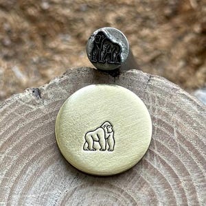 Op de afbeelding: Een metalen stempel en een gestempelde messing schijf met een gorilla-ontwerp. De stempel is zilverkleurig en de schijf is goudkleurig. De gorilla is afgebeeld in een gedetailleerde, gestileerde omtrek. De items worden op een houten oppervlak weergegeven.