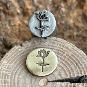Op de afbeelding: Twee ronde metalen stempels, één zilverkleurig en één goudkleurig, elk voorzien van een diep gegraveerd rozenontwerp. De stempels zijn verticaal geplaatst en tonen het gedetailleerde bloemmotief op hun platte, gepolijste oppervlakken.