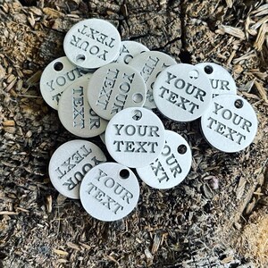 May include: A collection of silver-colored metal tags with the text "YOUR TEXT" engraved on each one. The tags have a hole punched in the top for attaching to a key chain or other item.