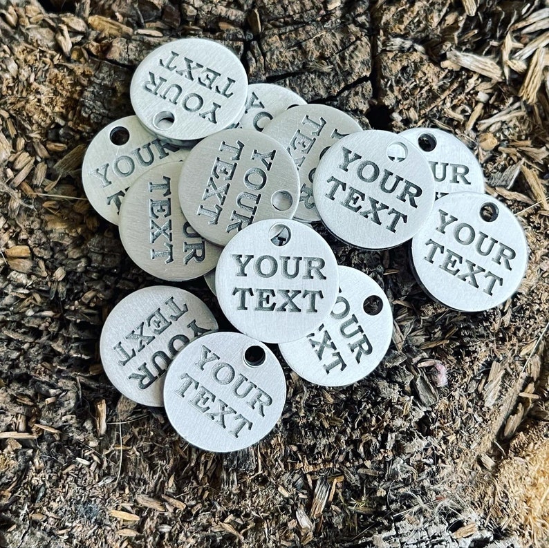 May include: A collection of silver-colored metal tags with the text "YOUR TEXT" engraved on each one. The tags have a hole punched in the top for attaching to a key chain or other item.