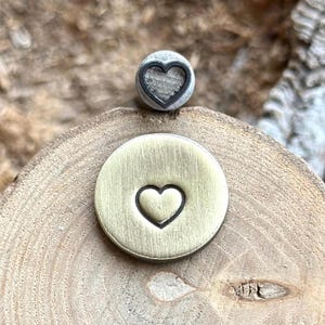 Può includere: Due timbri metallici con disegni a forma di cuore. Uno è un piccolo timbro rotondo color argento con un contorno a forma di cuore. L'altro è un timbro più grande, rotondo, color ottone con una forma di cuore premuta nel metallo.