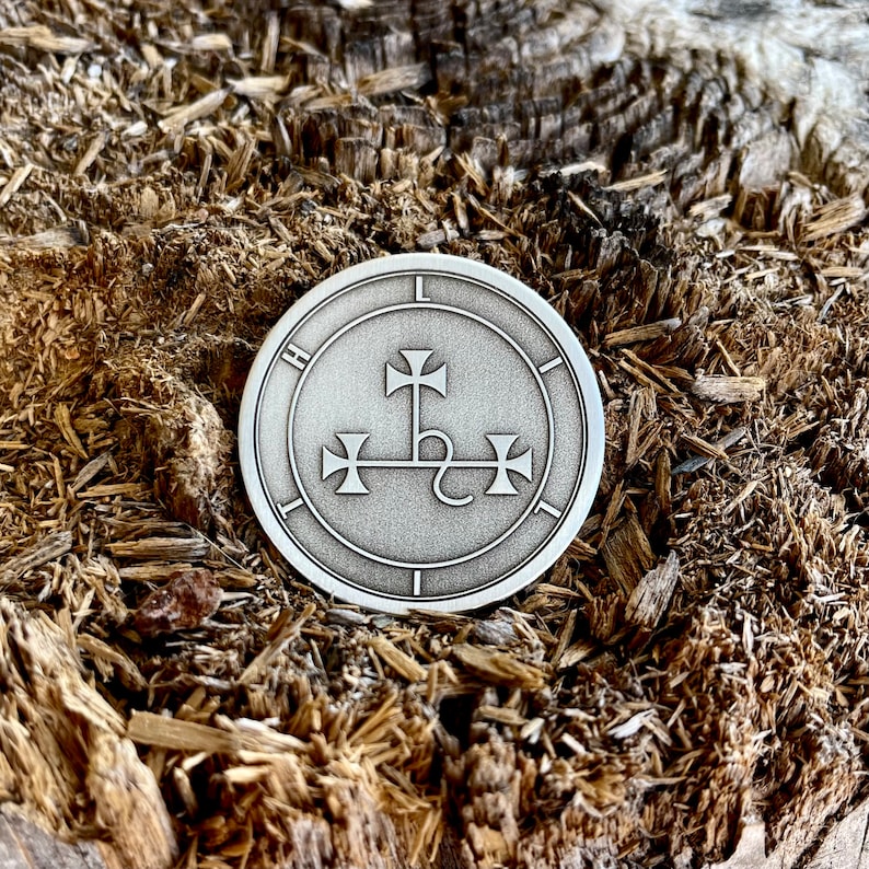 May include: A silver-colored round pendant with a detailed engraved design. The pendant features a central cross-like symbol with additional cross-like elements around the perimeter. The background is a textured, natural wood surface.