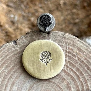 May include: A metal stamp and a stamped brass disc featuring a hydrangea flower design. The stamp is a small, round, metal tool with the flower design on its face. The disc is round and has the same flower design stamped into its surface.