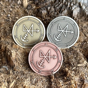 Puede incluir: Tres fichas metálicas con un diseño circular. Las fichas son de color dorado, plateado y cobre. Cada ficha presenta un símbolo con una flecha y un ancla. Las letras N, O, D, A, B y D están inscritas alrededor del borde de cada ficha.