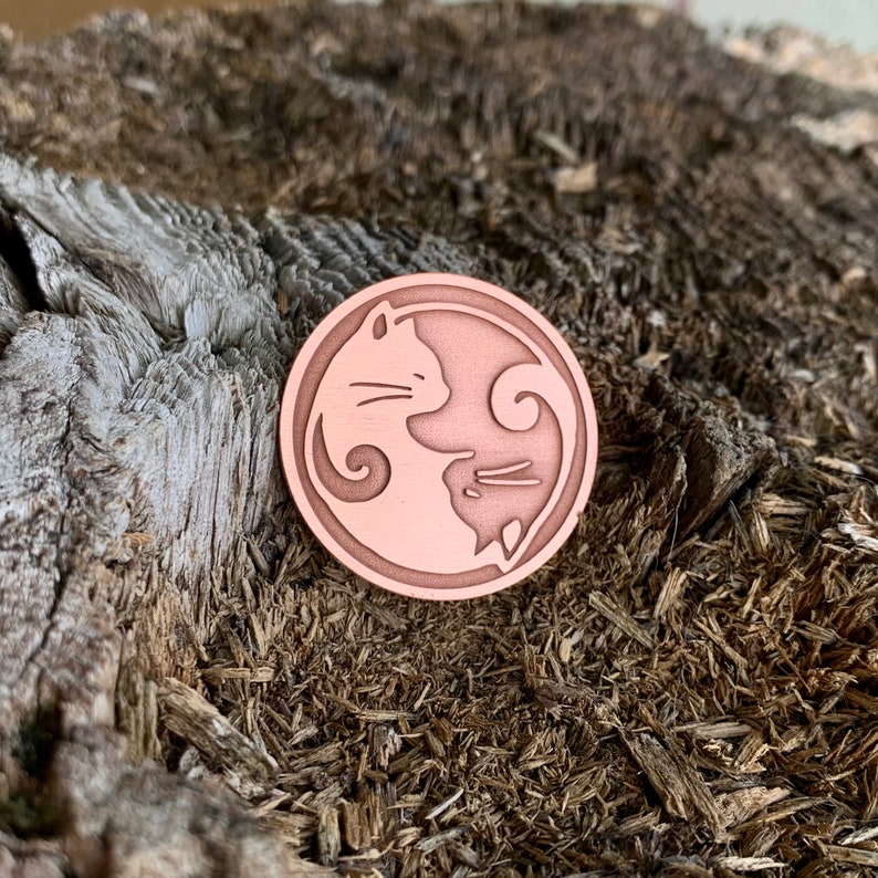 Op de afbeelding: Een koperkleurige pin met een yin-yang-ontwerp met twee katten die elkaar aankijken. De katten bevinden zich in een cirkelvorm met een swirl-ontwerp in het midden.