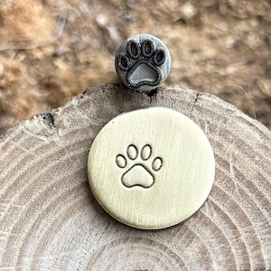 May include: A metal paw print stamp and a stamped impression on a round, gold-colored disc. The stamp is dark gray with a paw print design. The disc has a paw print impression.