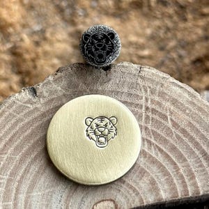 May include: A circular brass disc featuring a stamped tiger face design and a smaller, textured circular metal piece positioned above it, also displaying a stamped tiger face with a dark, oxidized finish and a detailed, rugged border.