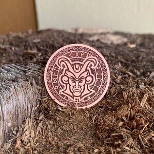 May include: A copper-colored coin with a detailed engraving of a horned, demonic face surrounded by a circular pattern of runes. The coin is resting on a piece of wood.