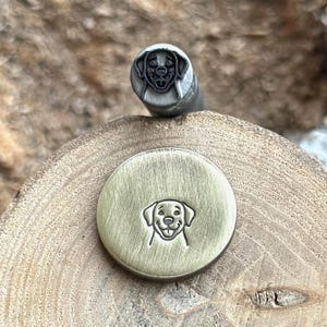 May include: A metal stamp featuring a Labrador Retriever dog face design. The stamp is made of metal and has a detailed engraving of the dog's features. A stamped metal disc with the same dog face design is also shown.