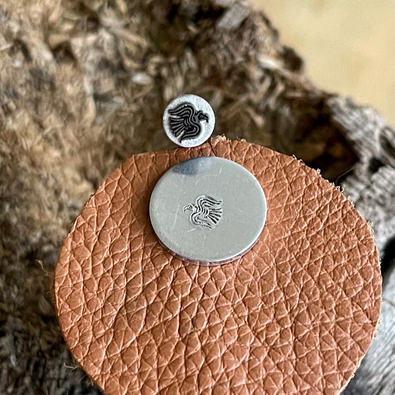 May include: Two silver metal buttons with a black and white bird design. One button is small and round, the other is larger and circular. They are placed on a brown leather surface.
