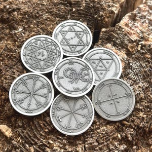 Puede incluir: Una colección de medallones circulares plateados con diseños y símbolos intrincados. Cada medallón presenta patrones únicos, incluyendo estrellas, formas geométricas y texto. Los medallones están dispuestos sobre una superficie de madera marrón texturizada.