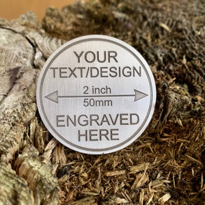 Könnte beinhalten: Eine runde silberne Metallplatte mit dem Text "YOUR TEXT/DESIGN" und "ENGRAVED HERE". Die Platte hat einen Durchmesser von 2 Zoll oder 50 mm.