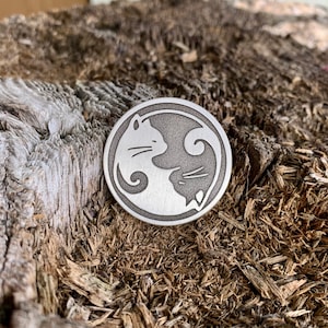 Op de afbeelding: Een zilveren metalen schijf met een yin yang-ontwerp met twee katten die elkaar aankijken. De katten zijn in een yin yang-ontwerp, met &eacute;&eacute;n kat aan de witte kant en de andere kat aan de zwarte kant.