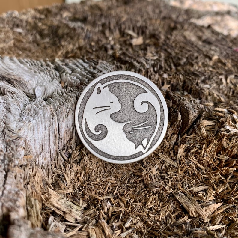 Op de afbeelding: Een zilveren metalen schijf met een yin yang-ontwerp met twee katten die elkaar aankijken. De katten zijn in een yin yang-ontwerp, met &eacute;&eacute;n kat aan de witte kant en de andere kat aan de zwarte kant.